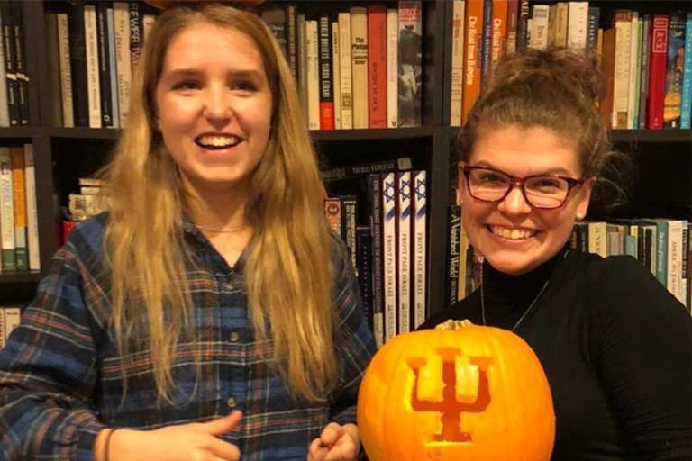 Two college students pose with carved pumpkin in library.